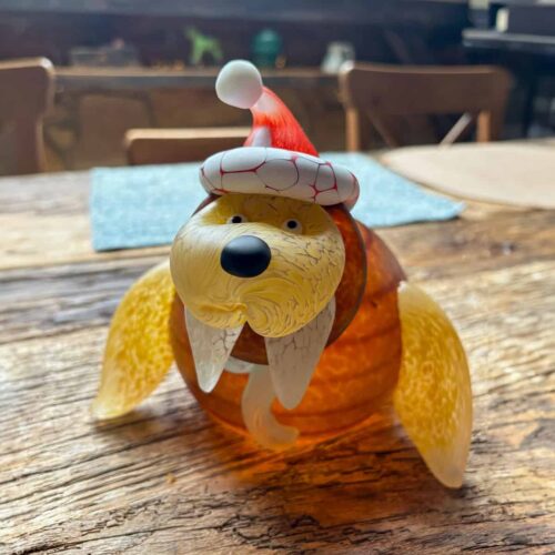 Glasfigur eines Walrosses mit Weihnachtsmütze auf einem Holztisch. Die Figur namens Wally trägt weiße Stoßzähne, einen Schal und strahlt festliche Gemütlichkeit aus.